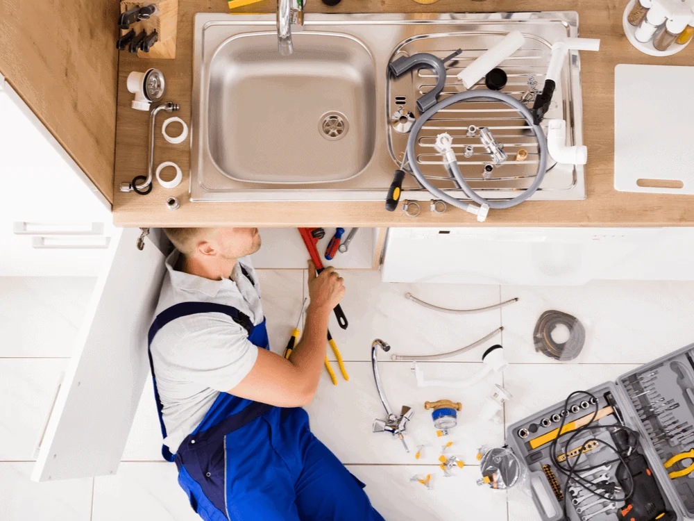 Plumber installing kitchen sink plumbing with tools and fittings laid out on the floor